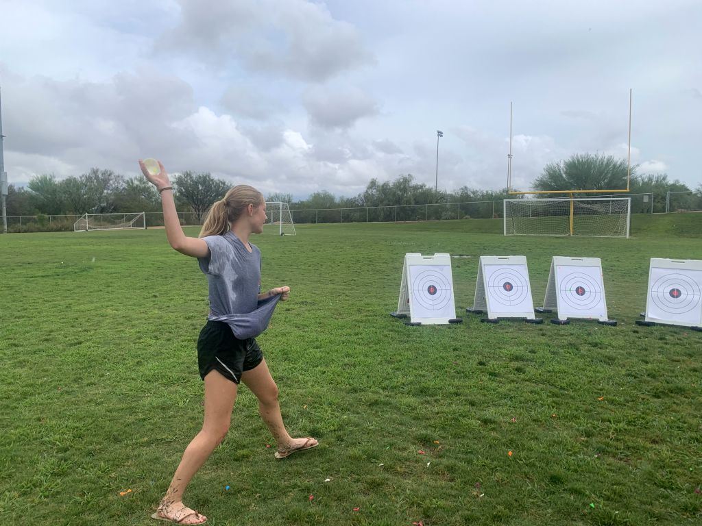 Teen girl preparing to toss a waterballoon at a row of targets on a soccer field.